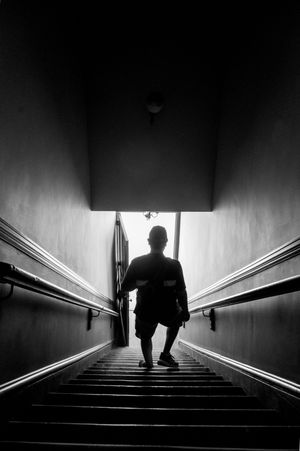 A dramatic black and white silhouette shows a person ascending a stairwell with handrails in low light conditions.