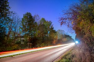 Light trails from cars streak through a dark road lined with autumn trees.