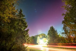 Long exposure night shot of car trails on road with colorful aurora sky and trees.