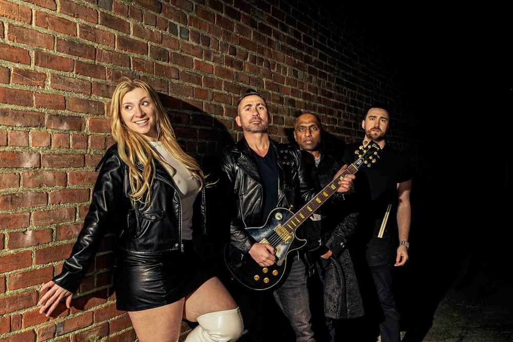 Musicians pose near a brick wall in dramatic lighting for a band photo.
