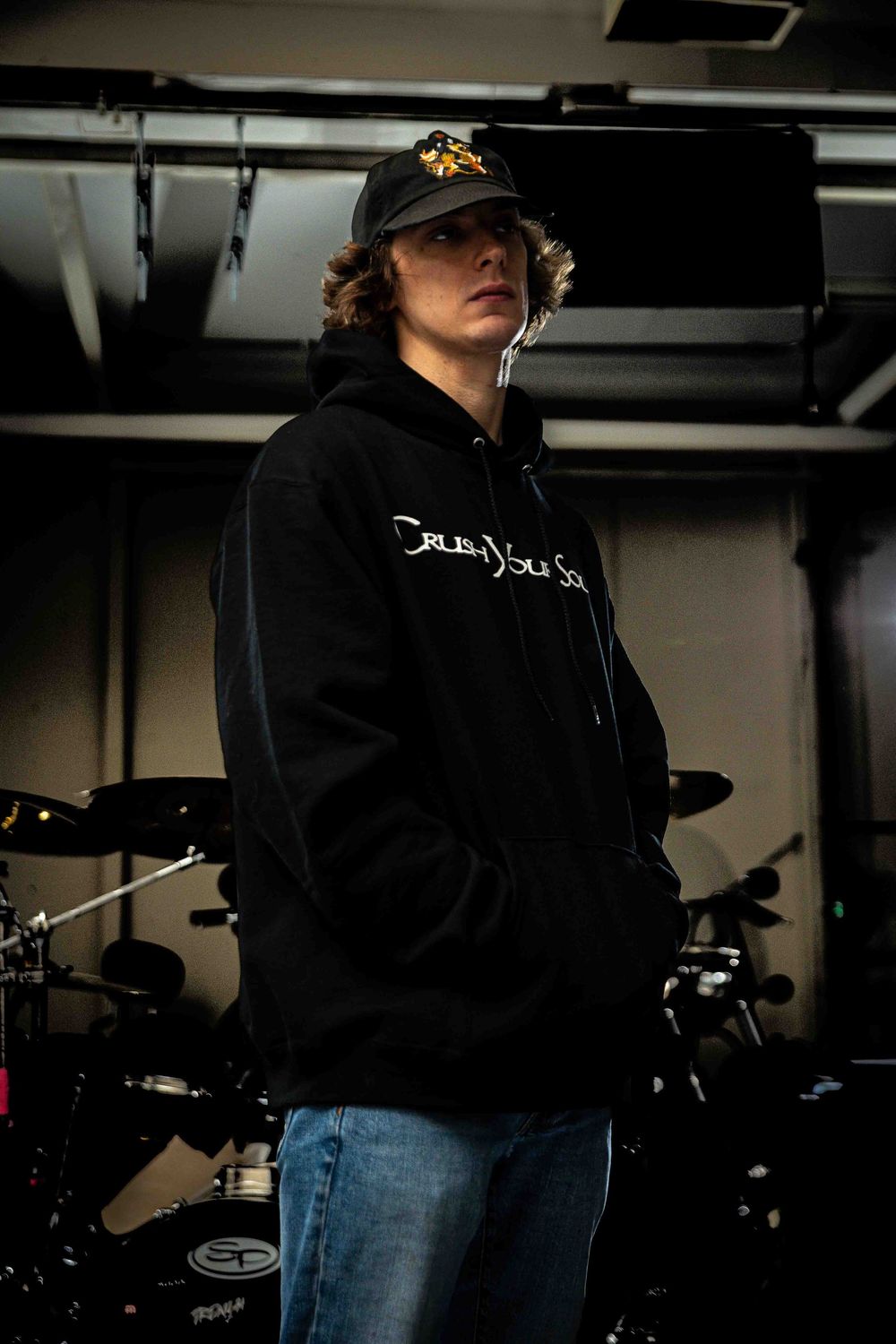 A musician in a black hoodie stands in a dimly lit rehearsal space.