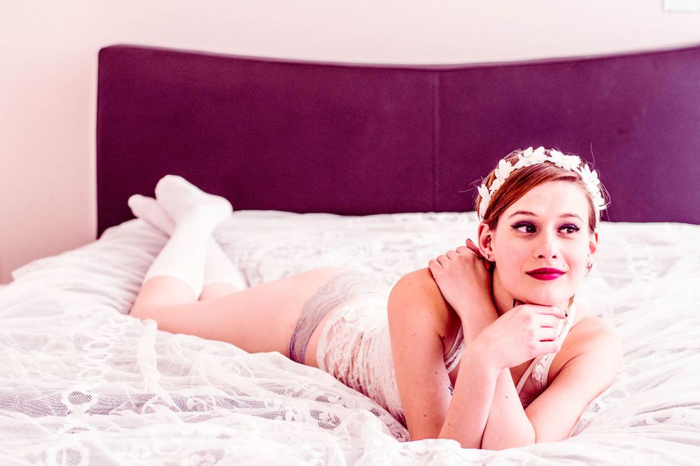 A figure in white lace lingerie reclines on a white bed with a jeweled headband against a purple wall.