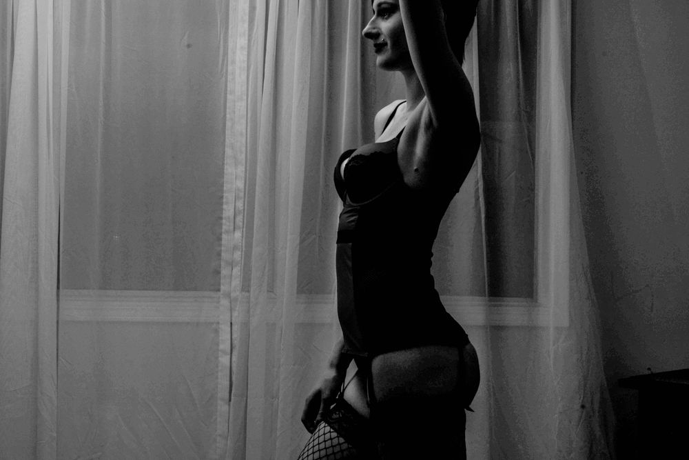Artistic black and white silhouette of a figure in lingerie posing dramatically by a window with sheer curtains.