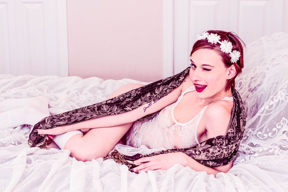 A vintage-style boudoir photo featuring delicate black lace, pearl necklace, and floral headpiece in soft pink tones.