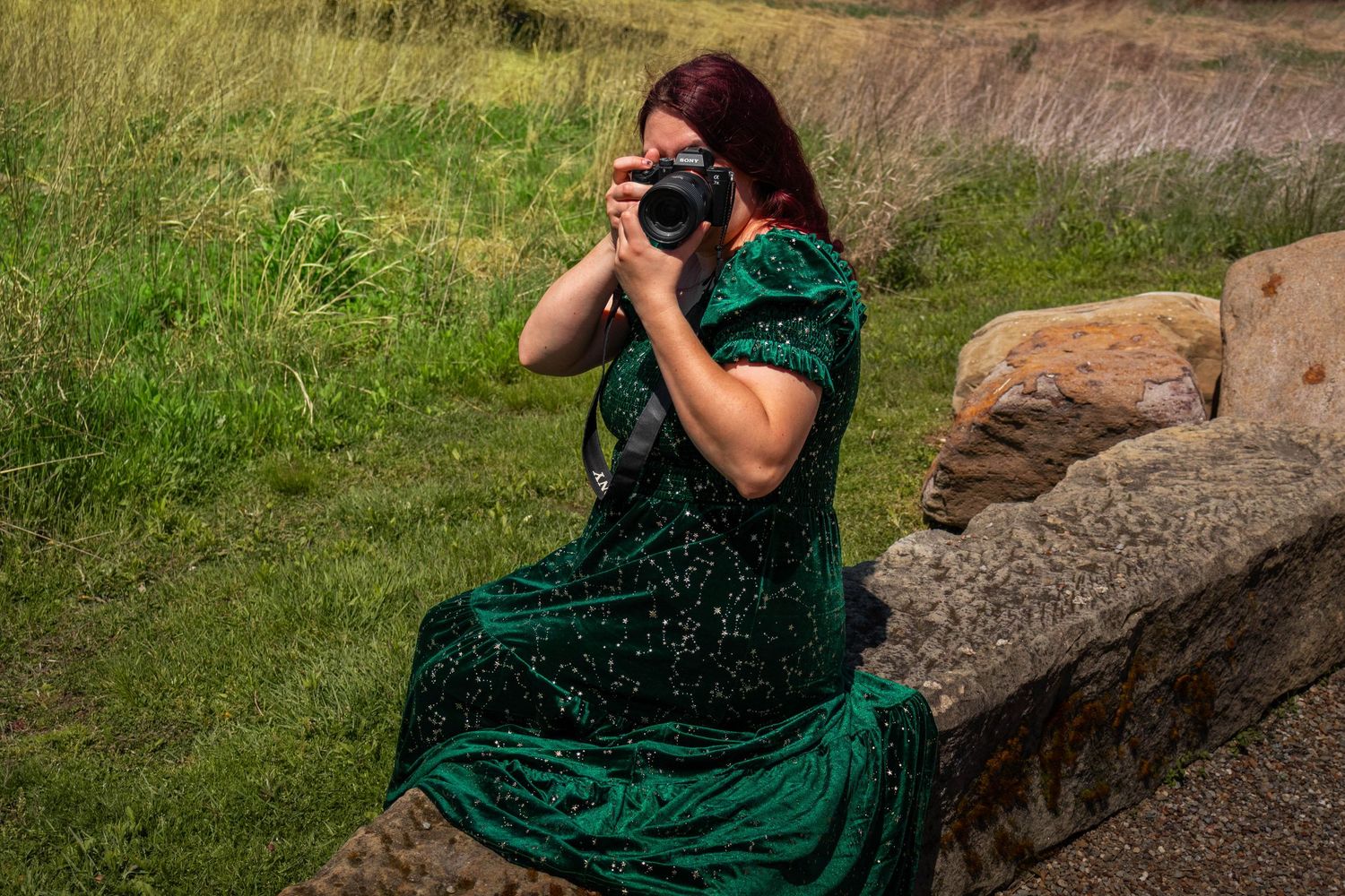 Red-haired woman in green velvet dress sitting on rocks photographing with DSLR camera.