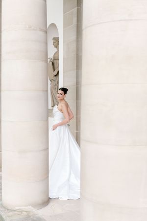 An elegant bride in a flowing white wedding gown poses gracefully between classic stone columns at a stately venue.