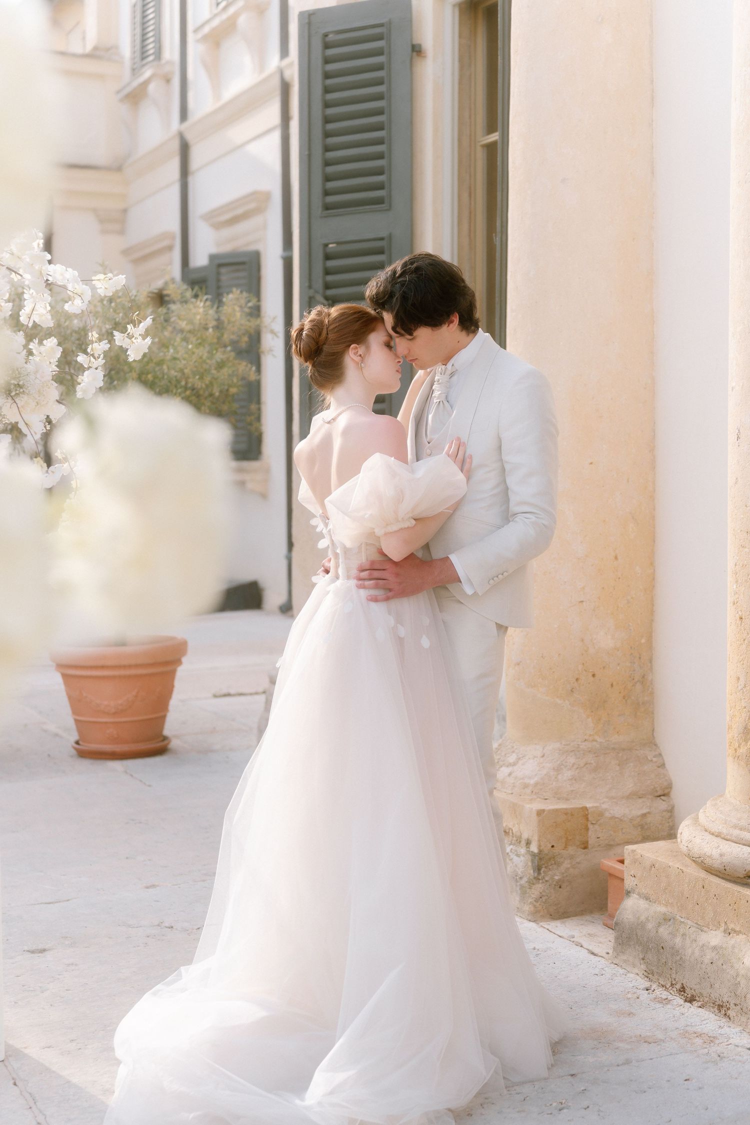 Romantic bridal couple in white wedding attire embrace against Italian villa exterior with stone columns and shutters.