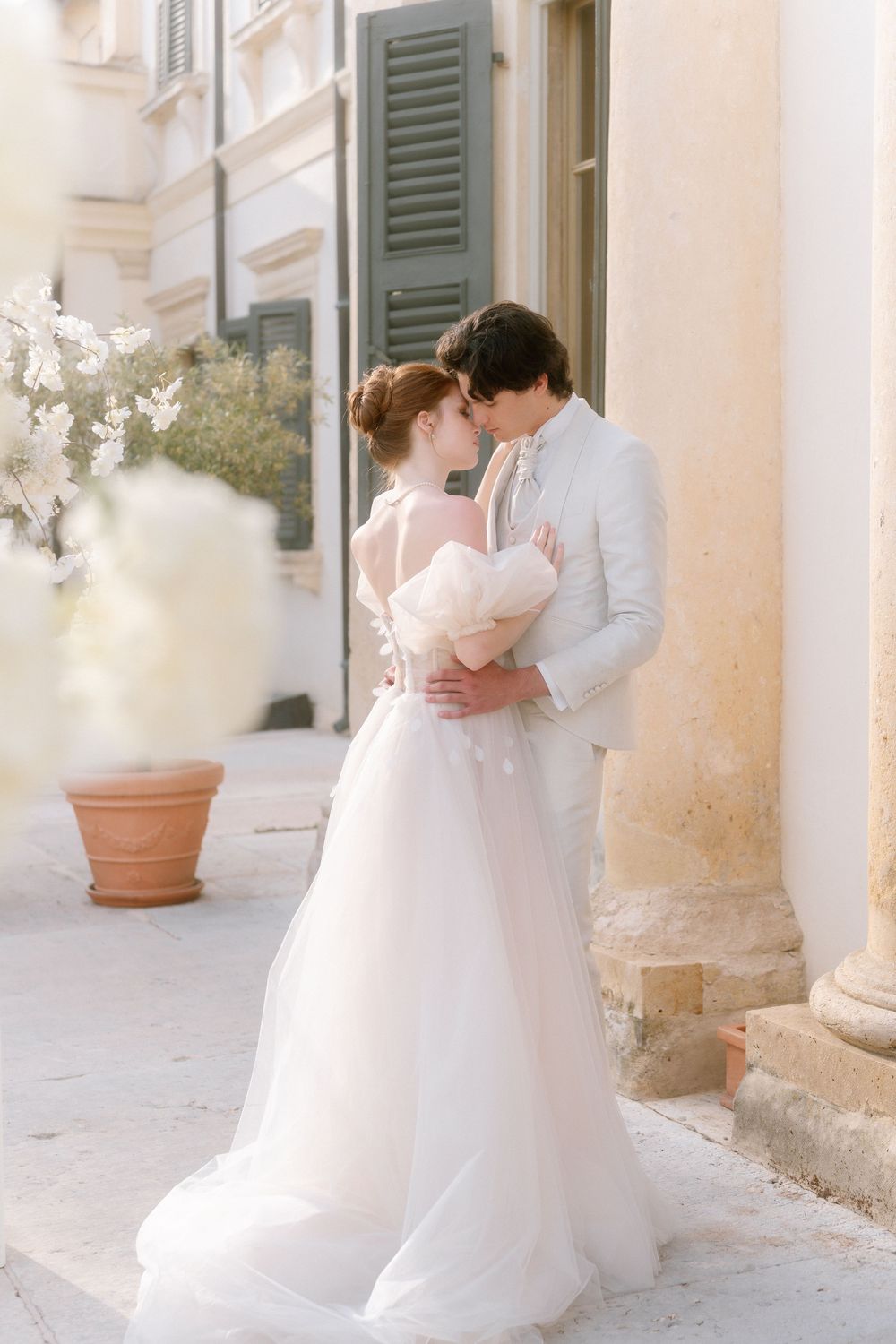 Romantic bridal couple in white wedding attire embrace against Italian villa exterior with stone columns and shutters.