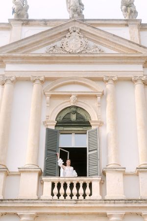 Figure in white garments appears at ornate balcony windows of historic white colonial building with classical columns.