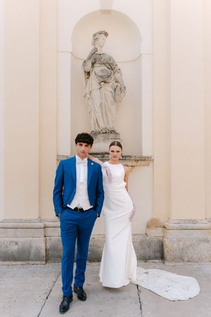 An elegant wedding couple poses against a classical stone wall with an ornate statue in an arched alcove above.