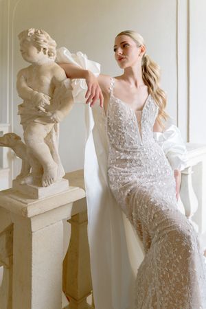 A white bridal gown with delicate lace detailing and plunging neckline displayed alongside classical marble statues.