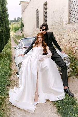 A couple poses with a vintage car against a rustic building wall on a scenic countryside path.