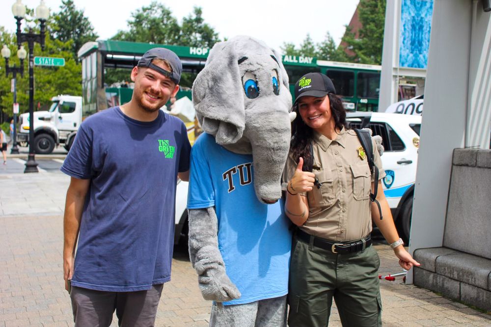 Two people pose with a mascot wearing a blue jersey outside a building.