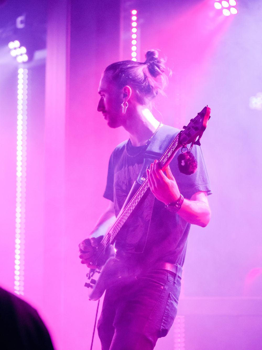 Musician with hair in bun playing bass guitar under pink stage lights.