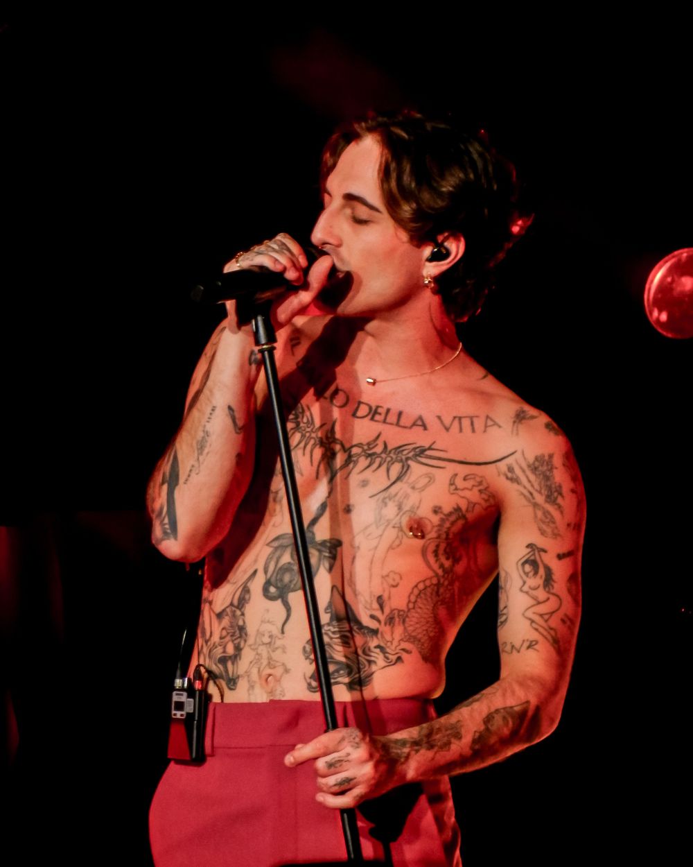 A performer in a red tank top sings passionately into a microphone on a dark stage with visible tattoos on their arms.