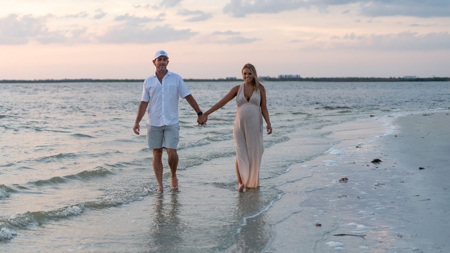 A couple walks hand in hand along the shoreline at sunset with calm ocean waters beside them.