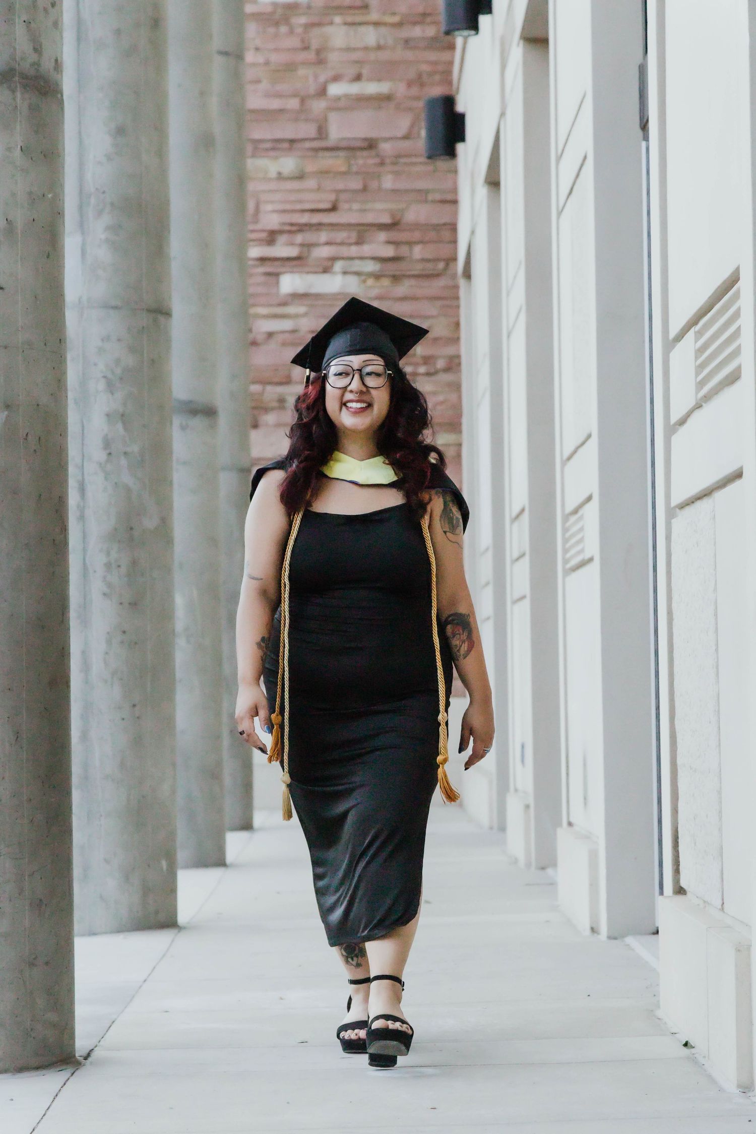 Graduate in black dress and cap walks through modern building colonnade wearing honor cords and sandals.
