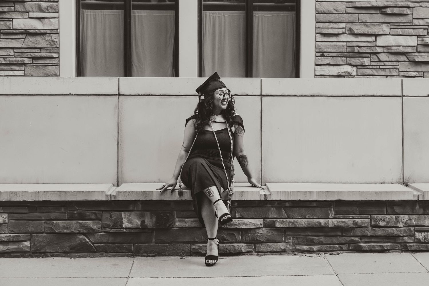 A person in a graduation cap and gown sits casually on a stone ledge outside a building in black and white.
