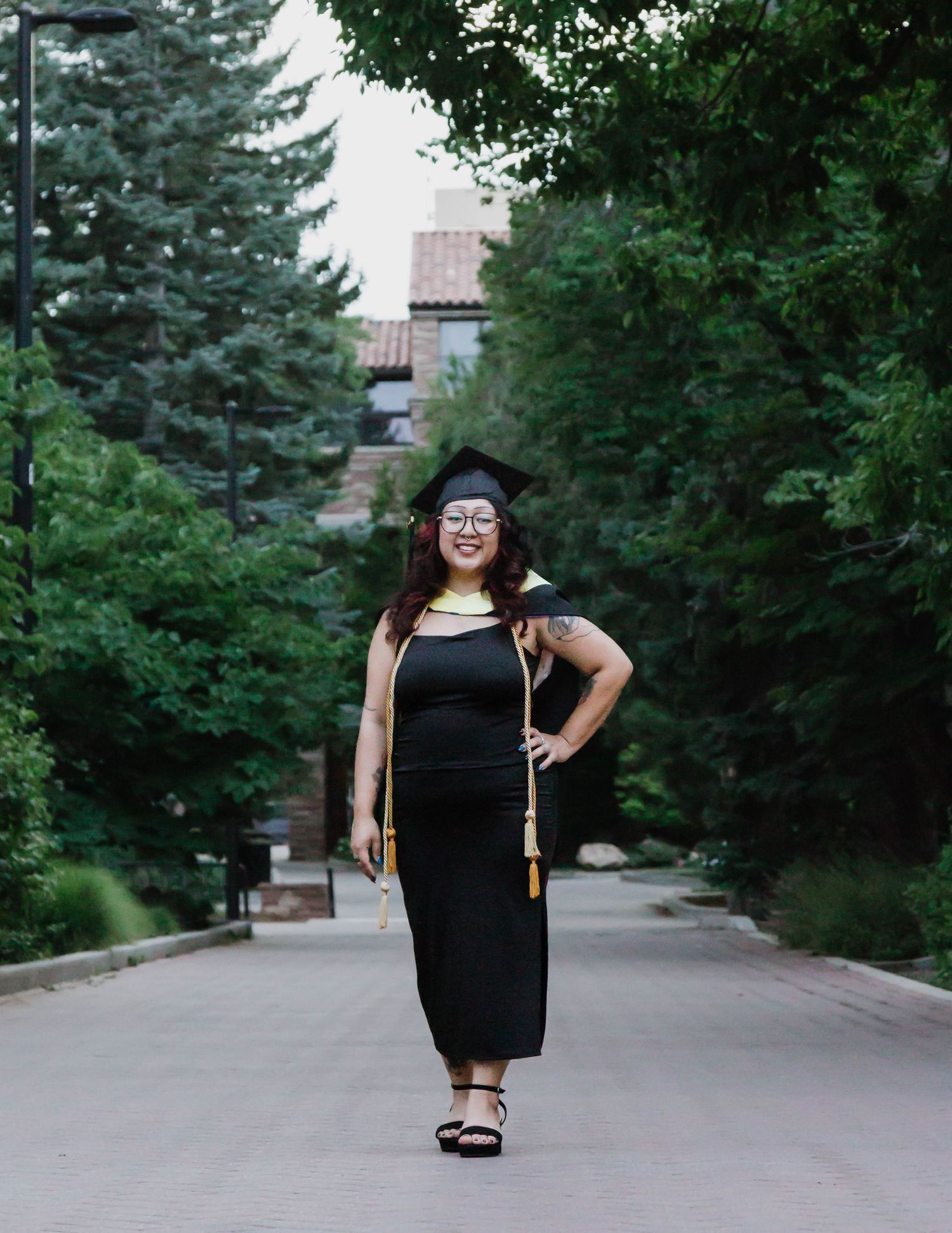 Someone wearing a black dress and graduation cap poses on a tree-lined pathway during a graduation photoshoot.