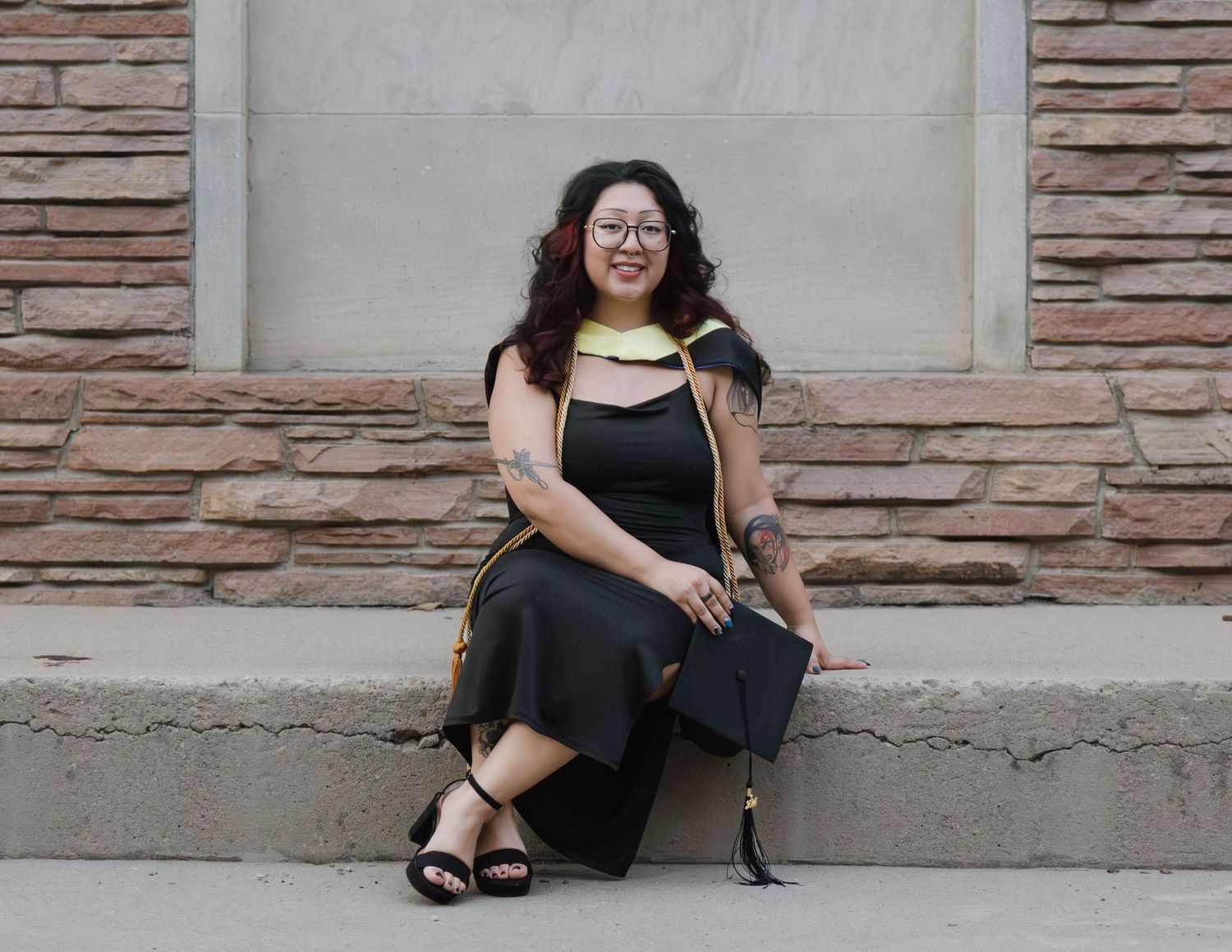 Someone in a black dress and yellow scarf sits smiling against a stone wall with a purse nearby.