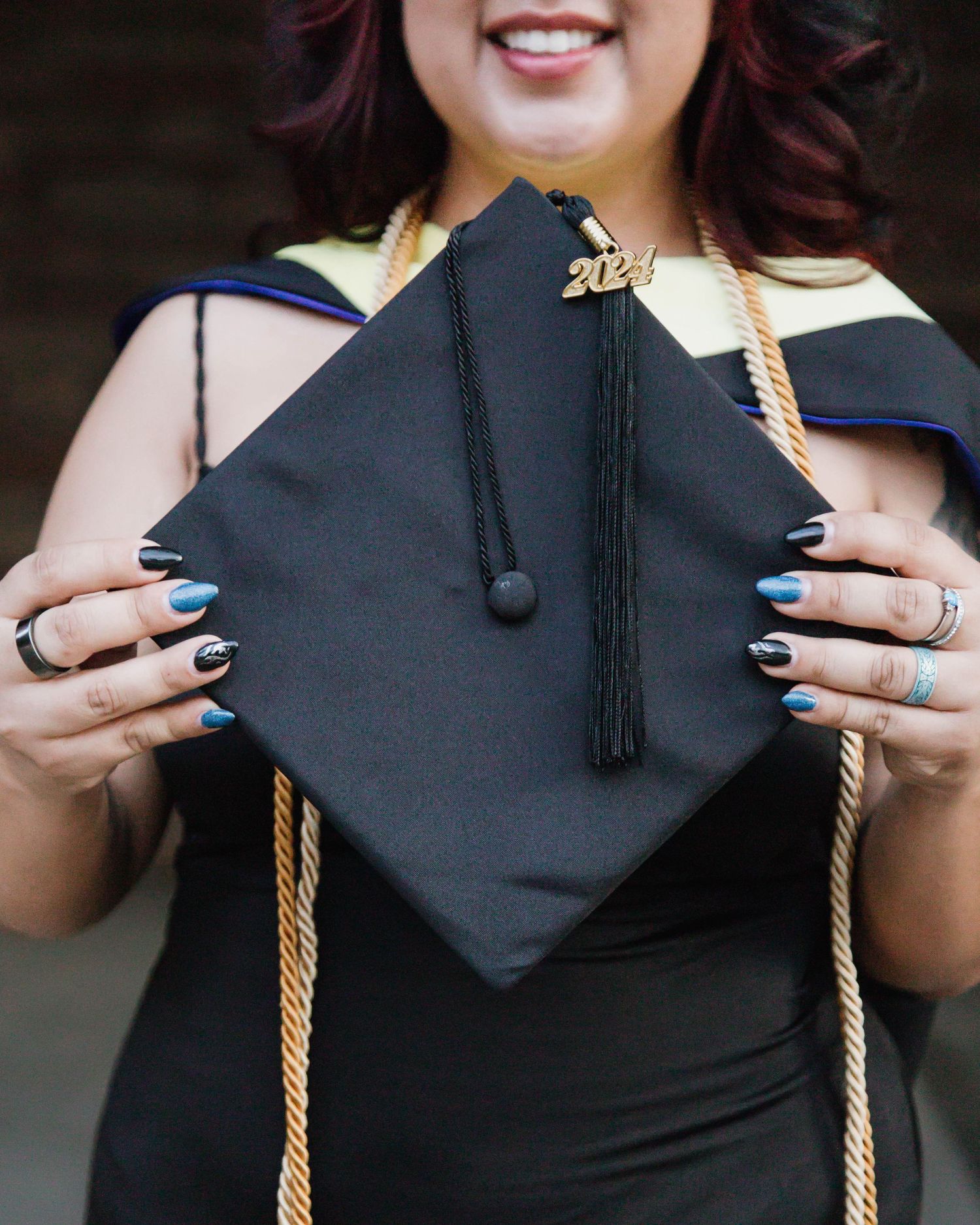 Graduate holding a decorated black graduation cap with blue nails and gold tassel cord against dark background.