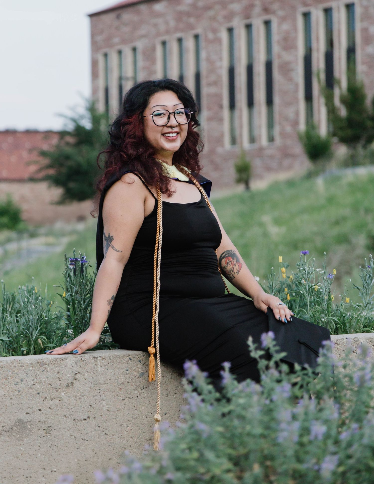 A person in a black dress and glasses sits on a stone wall surrounded by purple wildflowers near a brick building.