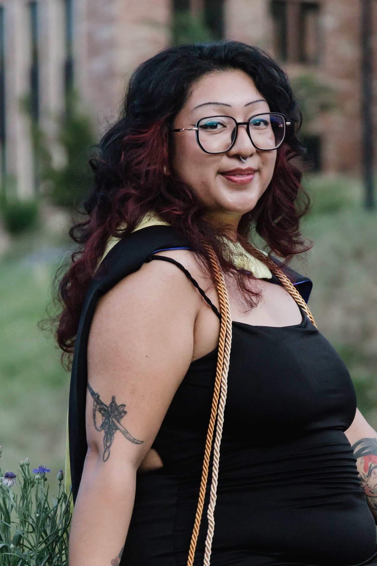 Graduate in black dress with burgundy hair and glasses poses outdoors near purple flowers with a gold honor cord.