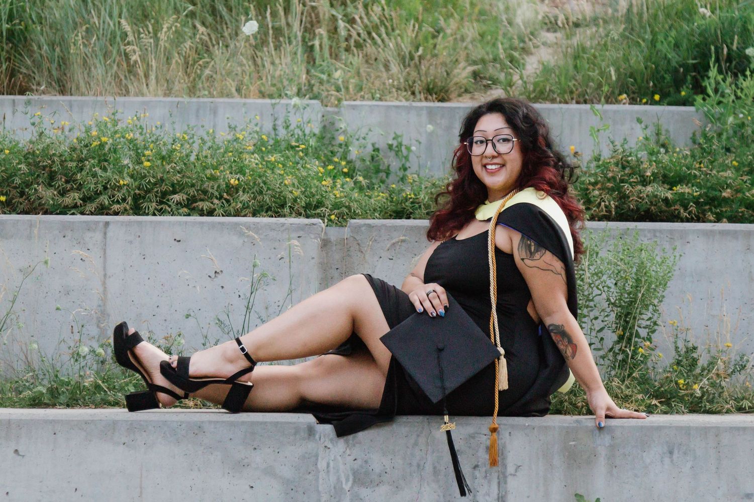 Graduate in black dress and graduation stole reclines on concrete steps during outdoor photo session.