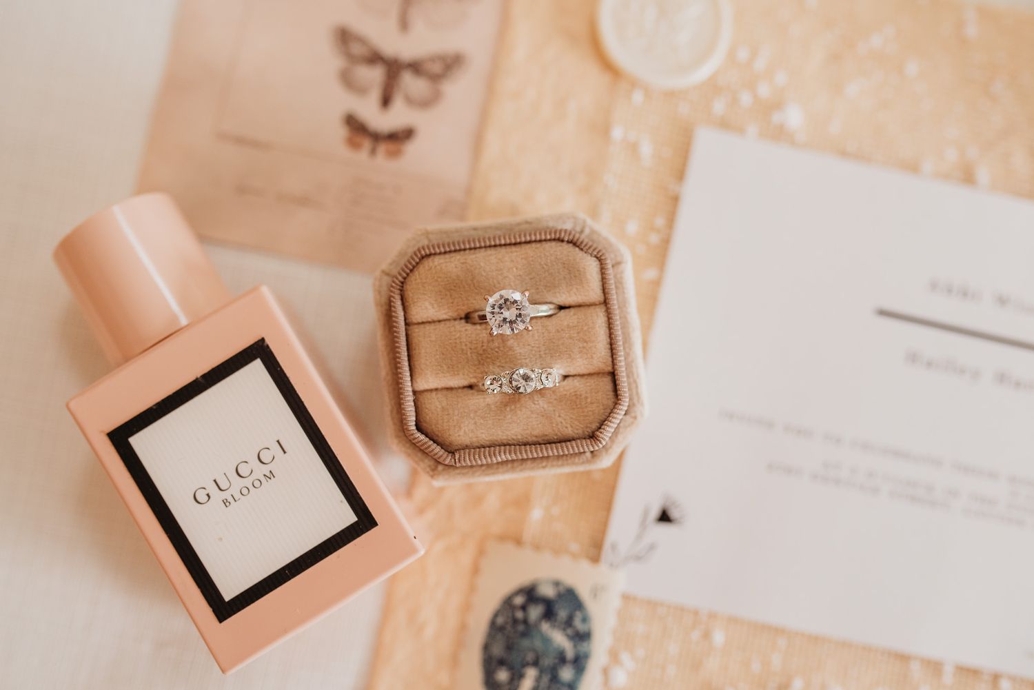Wedding details including pink perfume bottle, velvet ring box and invitation on beige surface.