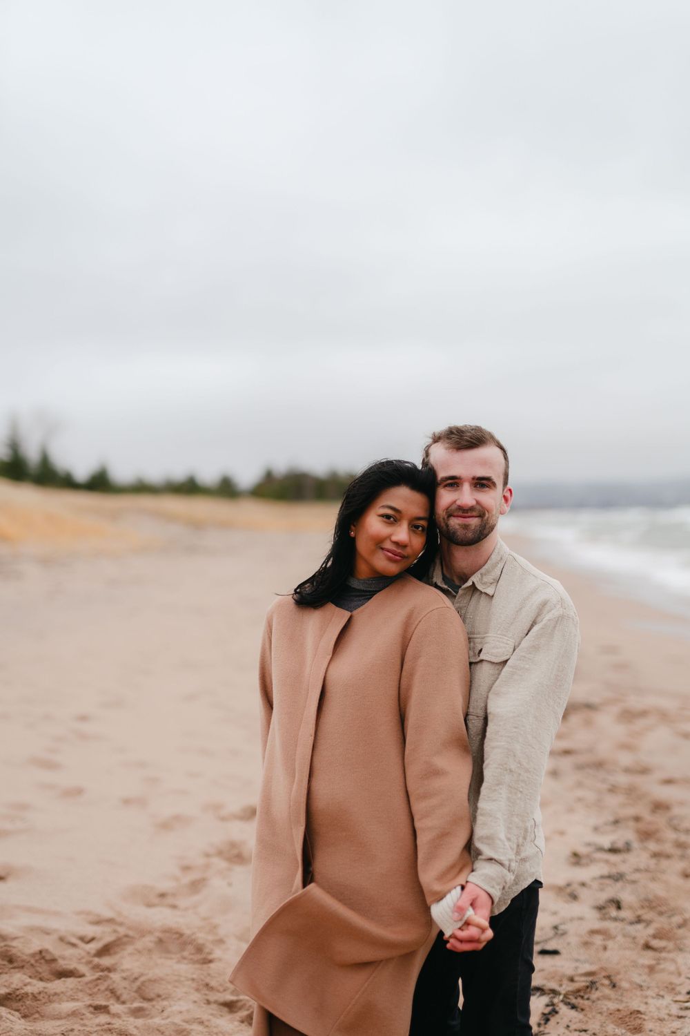 A couple poses together on a sandy beach on an overcast day.