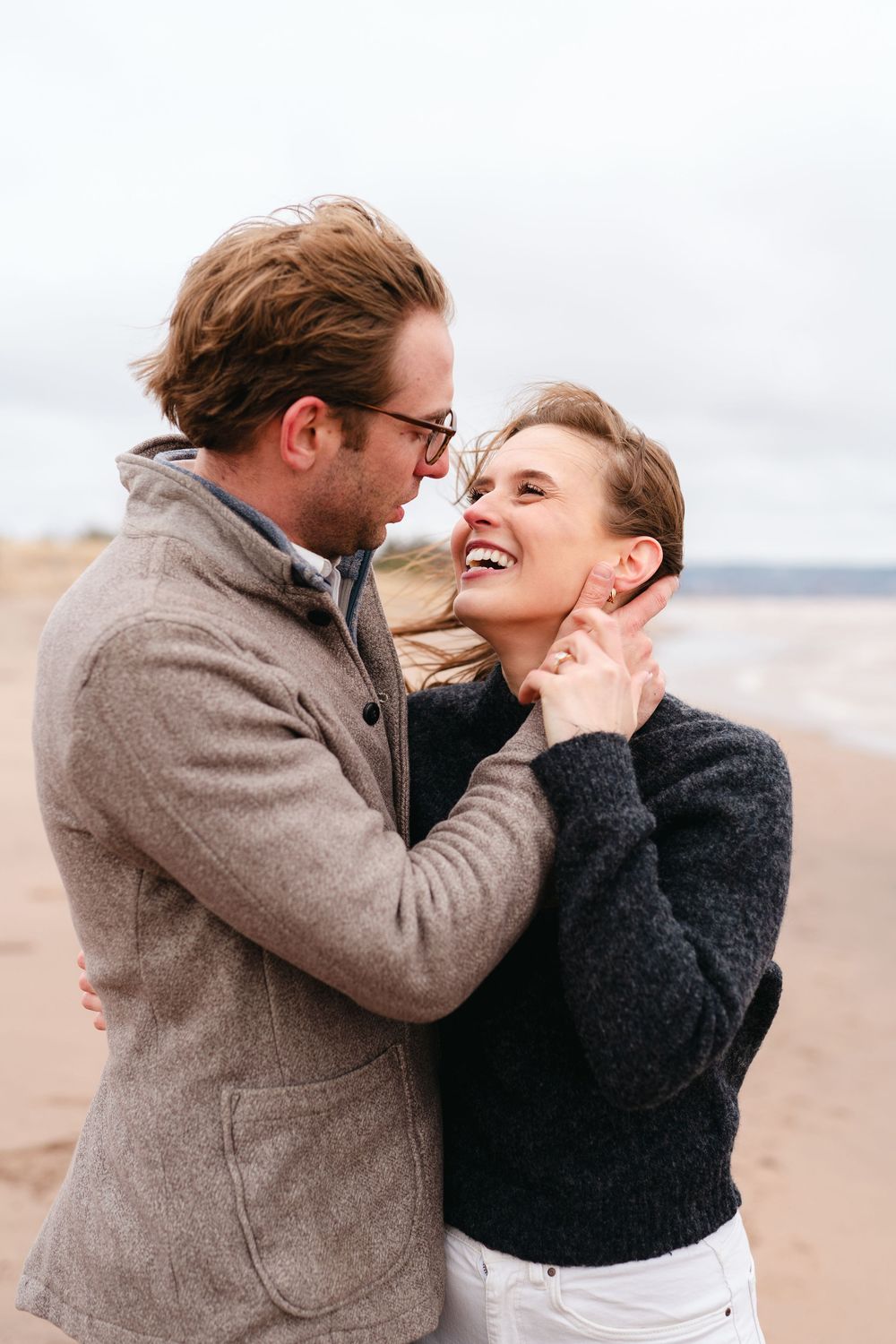 A romantic couple shares tender moments and laughter together on a cloudy beach while wearing cozy winter coats.