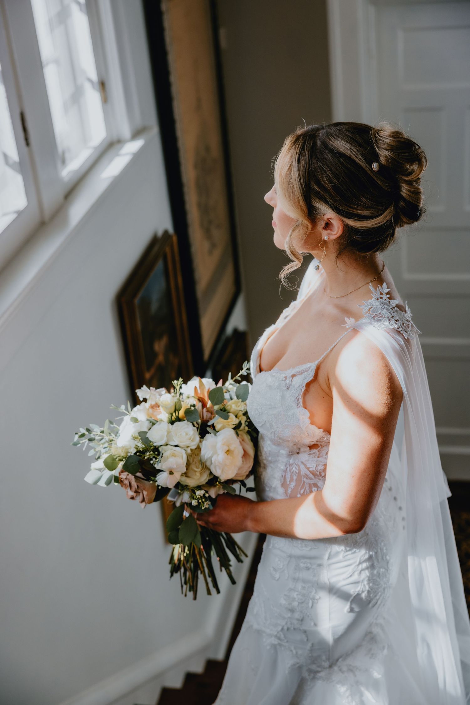 A bride in a lace wedding dress holds a beautiful white and cream floral bouquet while looking out a window.