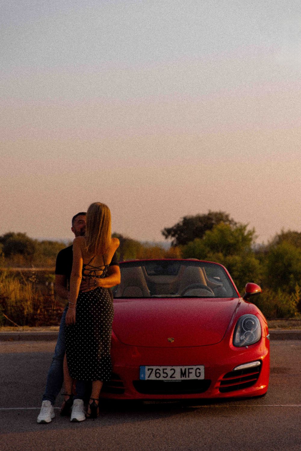 Person leaning against a red Porsche sports car at sunset.