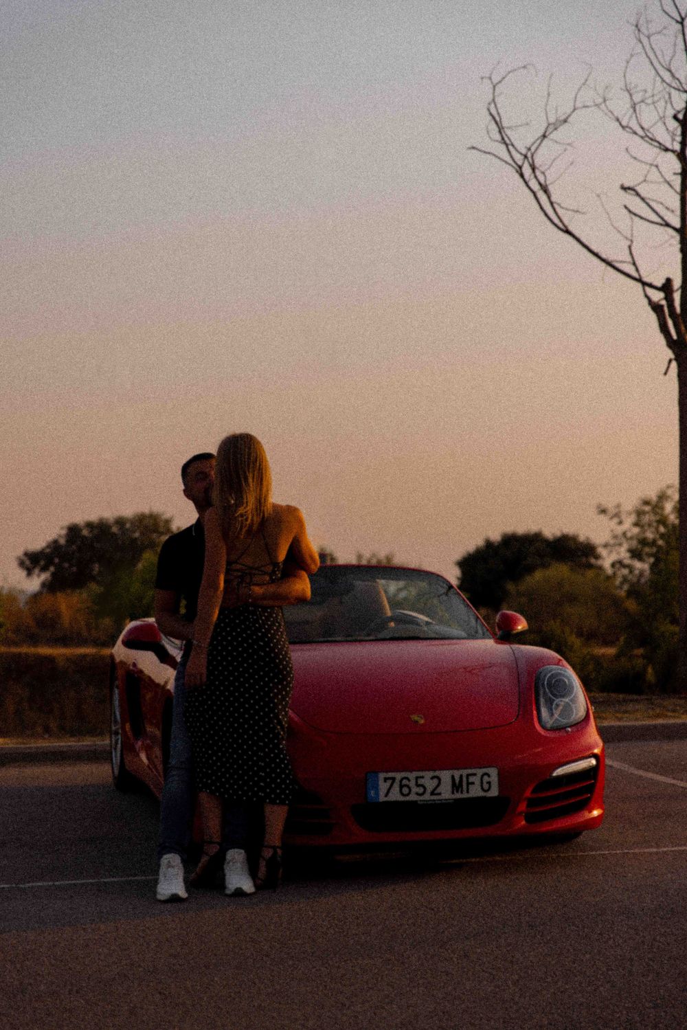 Person in polka dot dress poses beside red Porsche during sunset.