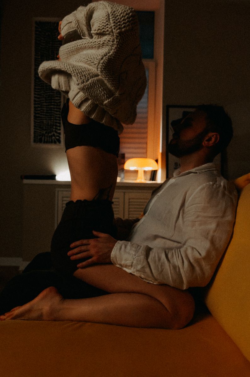 Intimate moody indoor scene with person in sweater and boots sitting on yellow couch in dimly lit room.