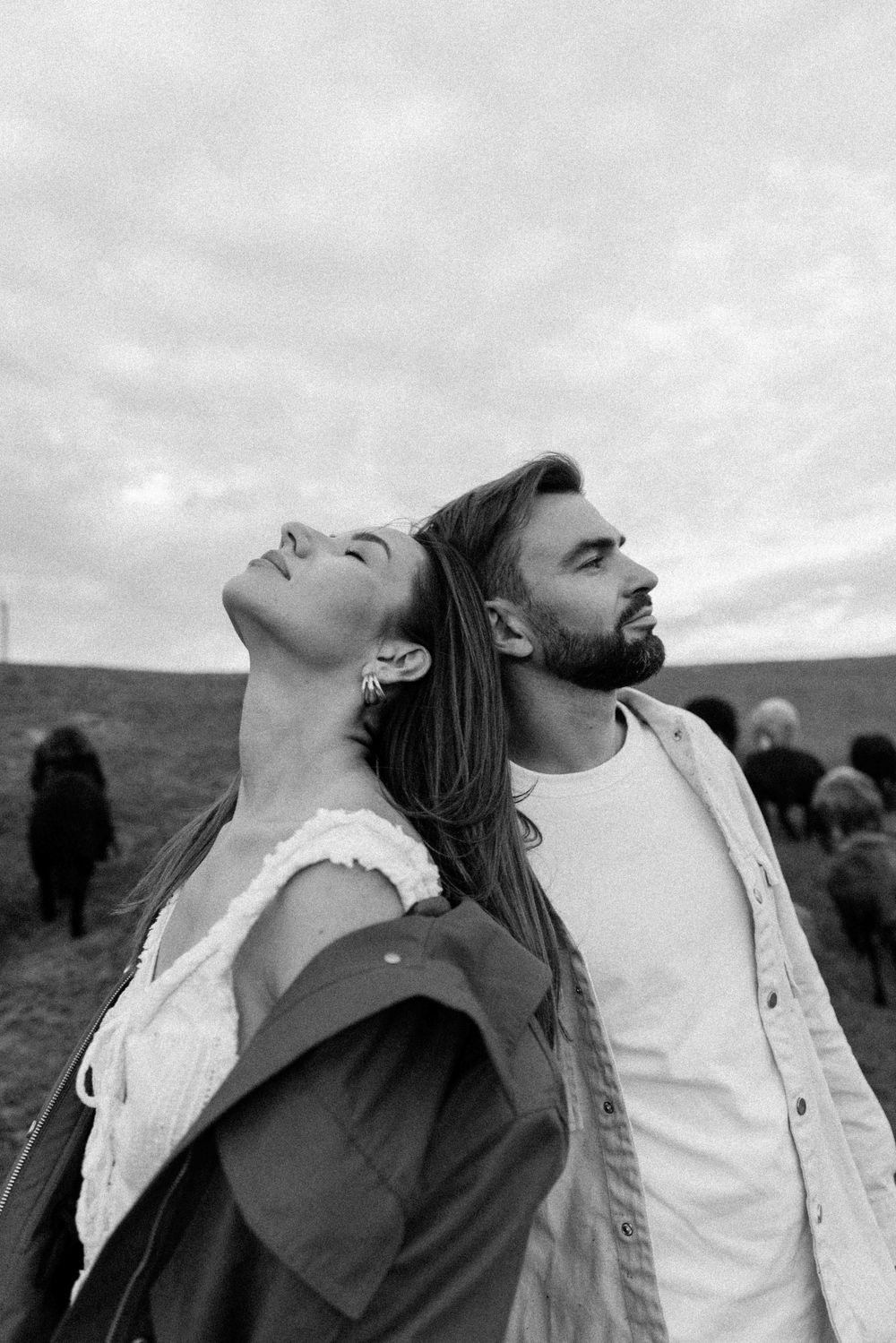 A couple in casual attire stands outdoors looking up at a cloudy sky in dramatic black and white photography.