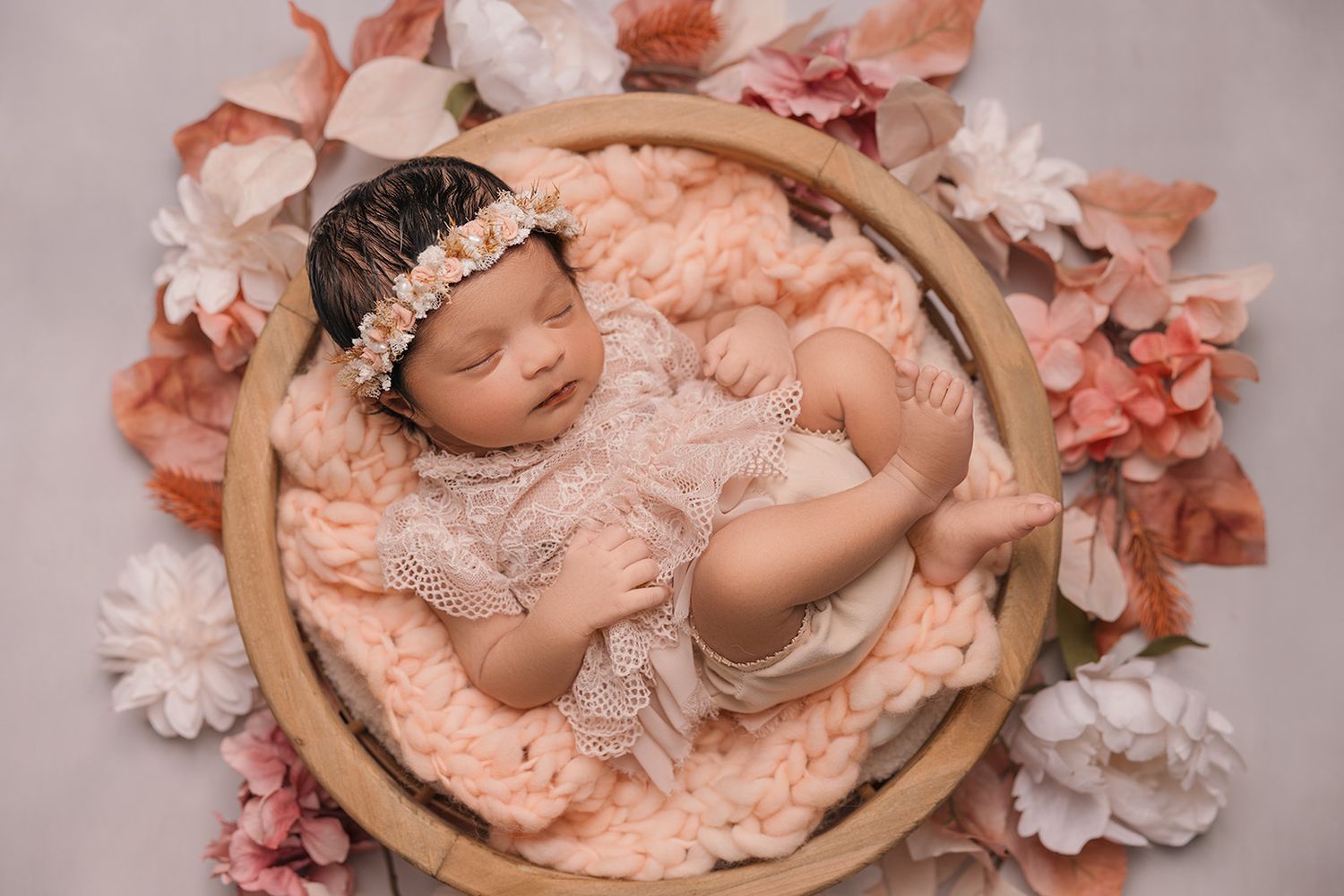 A sleeping newborn in lace wrap nestled in wooden bowl surrounded by pink and coral flowers.