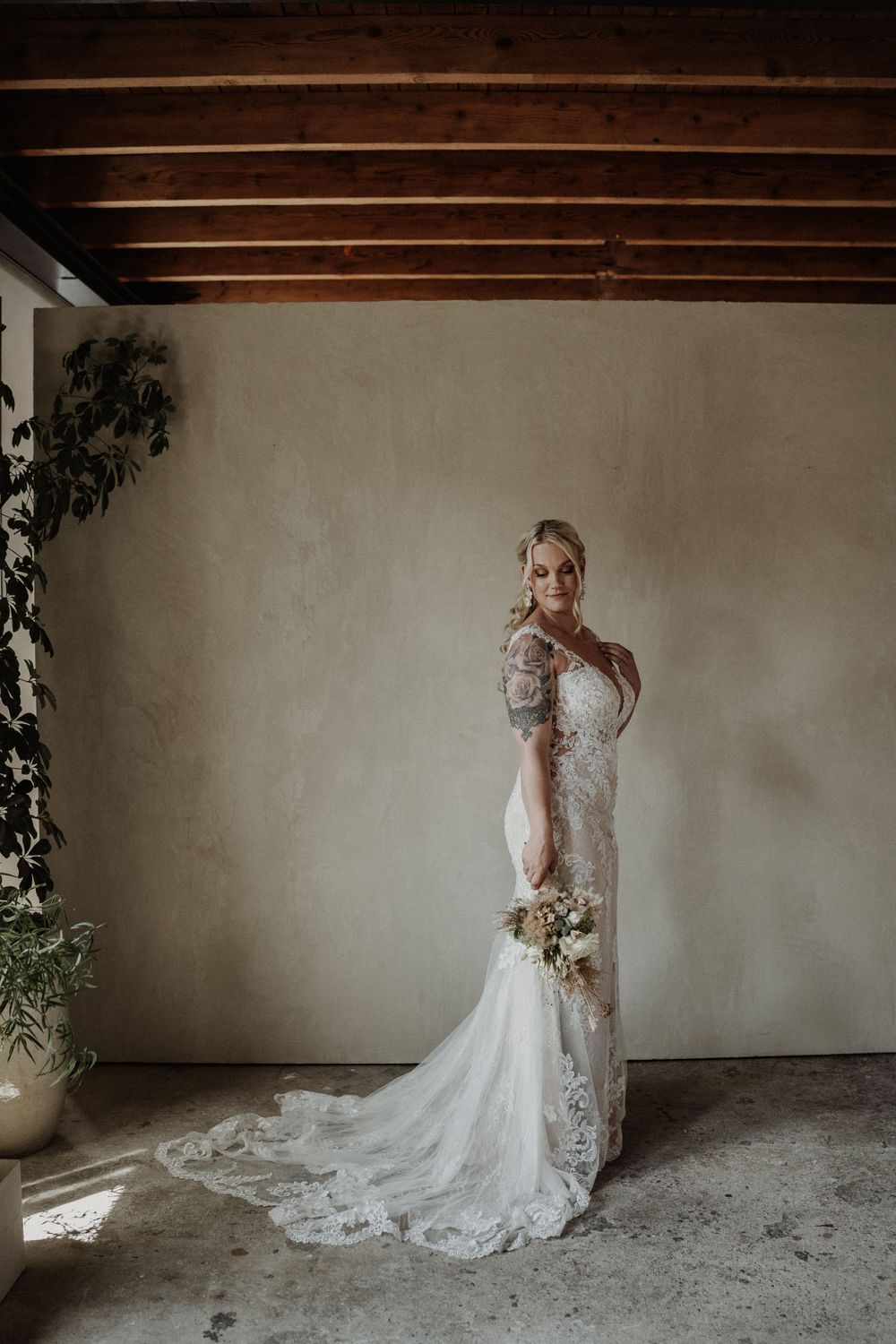 A bride in a fitted lace wedding gown with long train poses elegantly against a neutral wall under wooden beams.