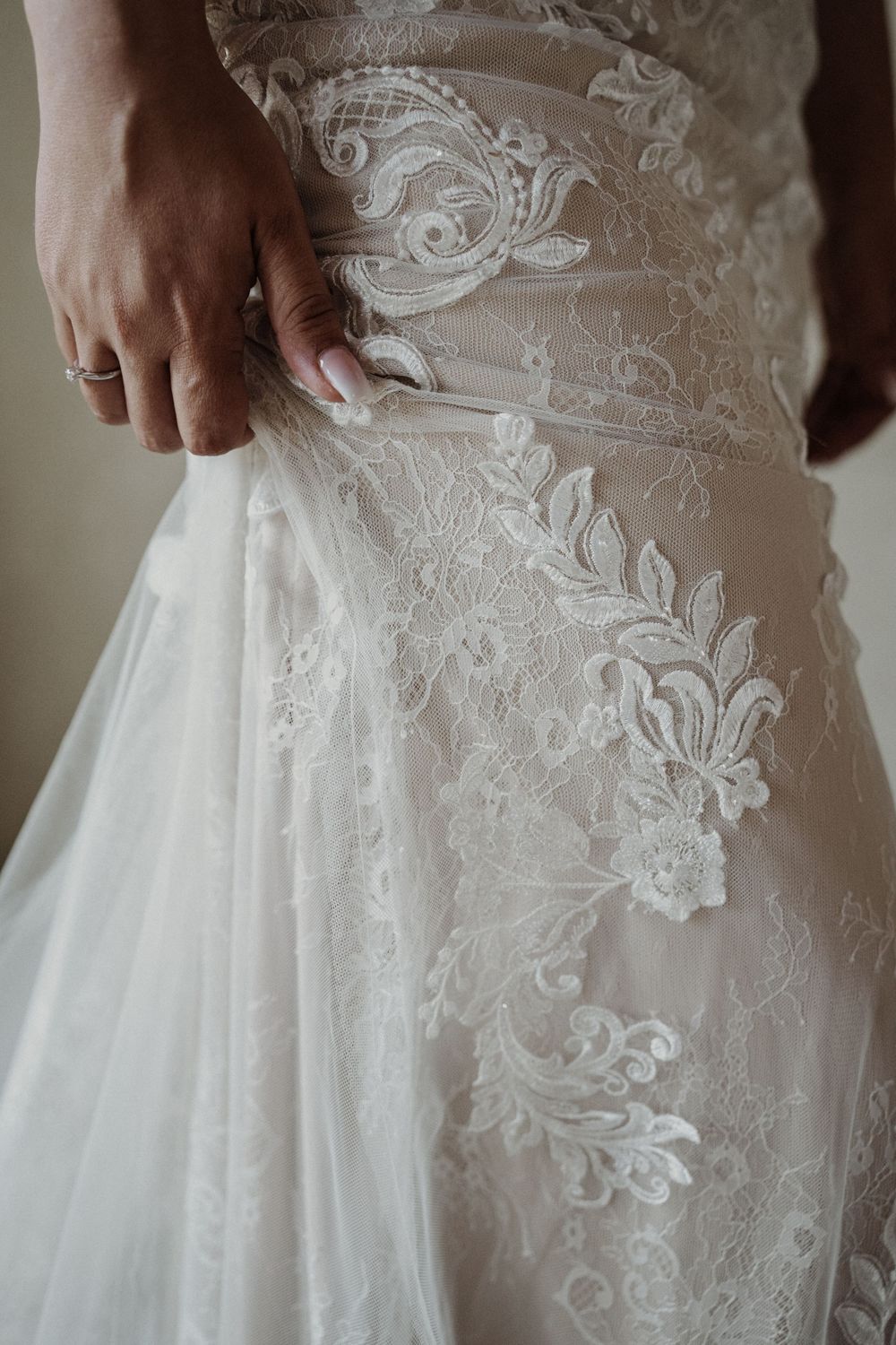 Close-up detail of delicate white lace and floral embroidery on a wedding dress with hand gently touching the fabric.