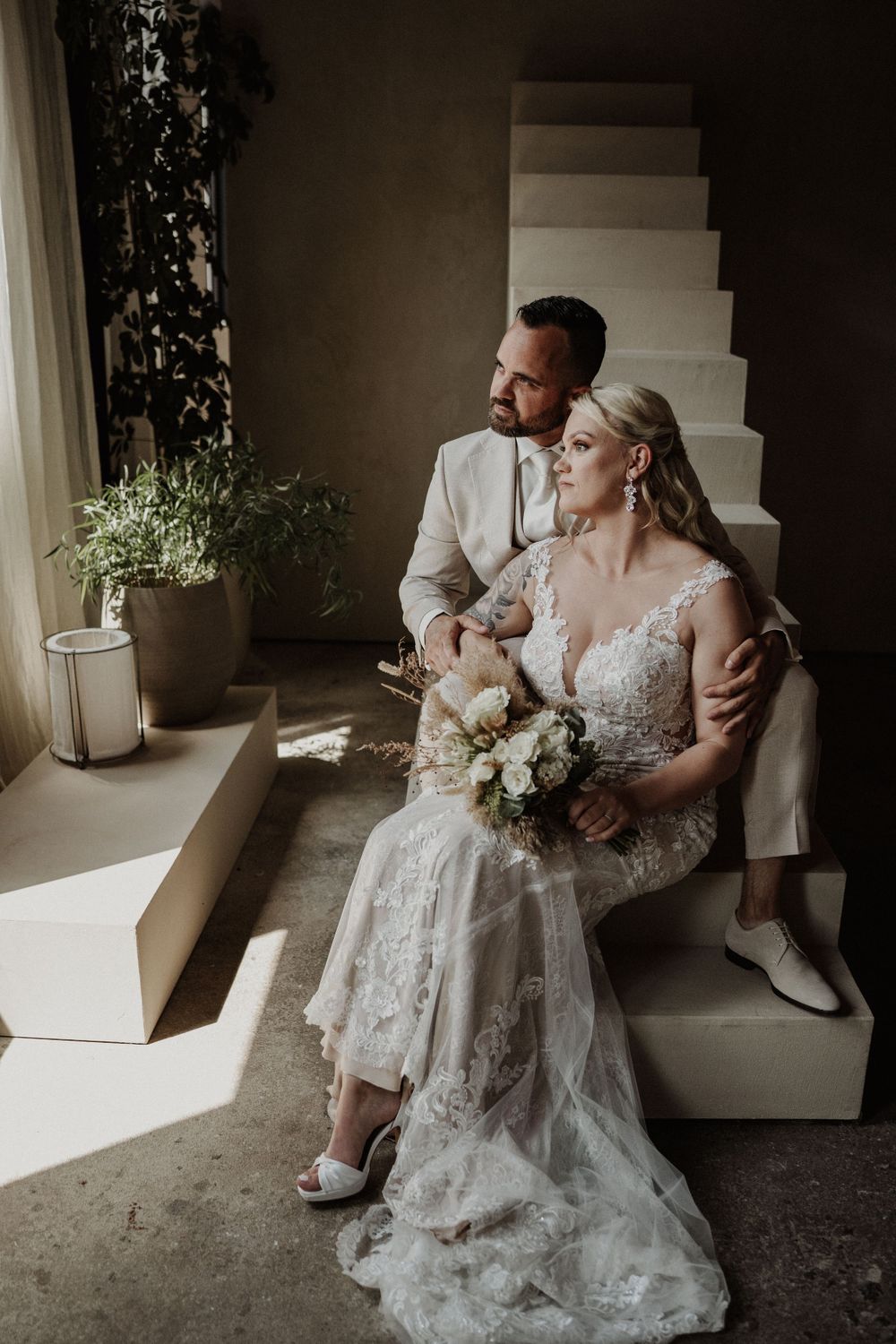 A couple sits together on light-colored steps, with the bride wearing a detailed lace gown and holding a white bouquet.