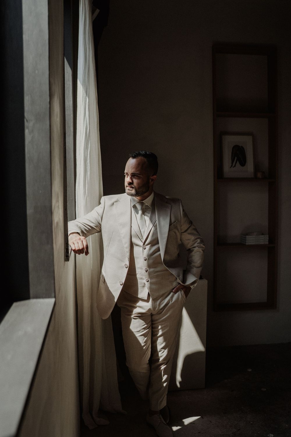 A groom in a cream-colored three-piece suit gazes thoughtfully out a window in moody natural lighting.
