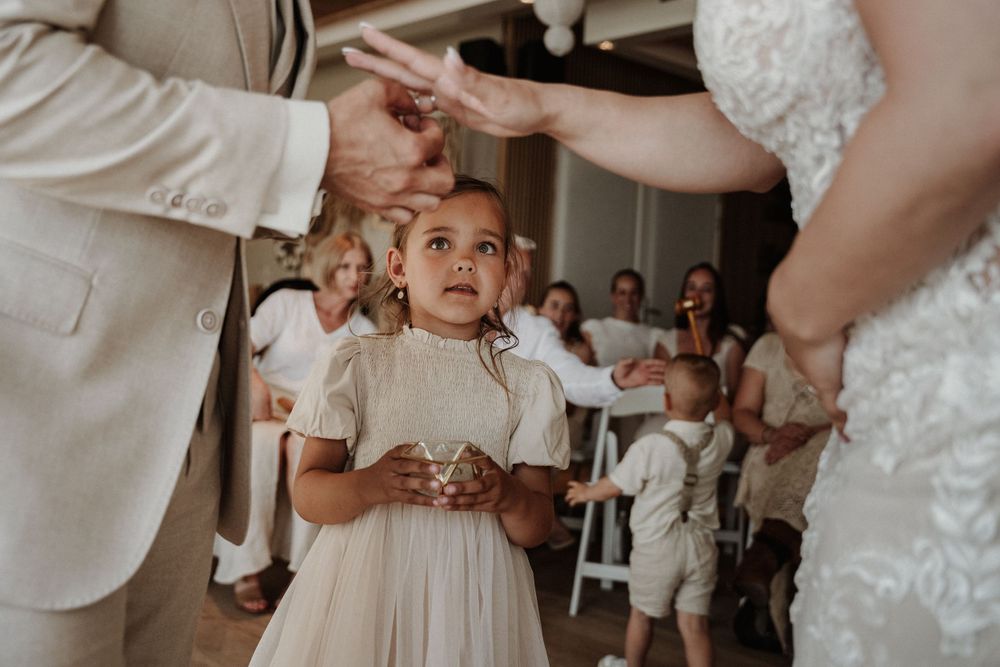 A small child in a white dress watches a wedding ceremony from the aisle while guests look on in the background.