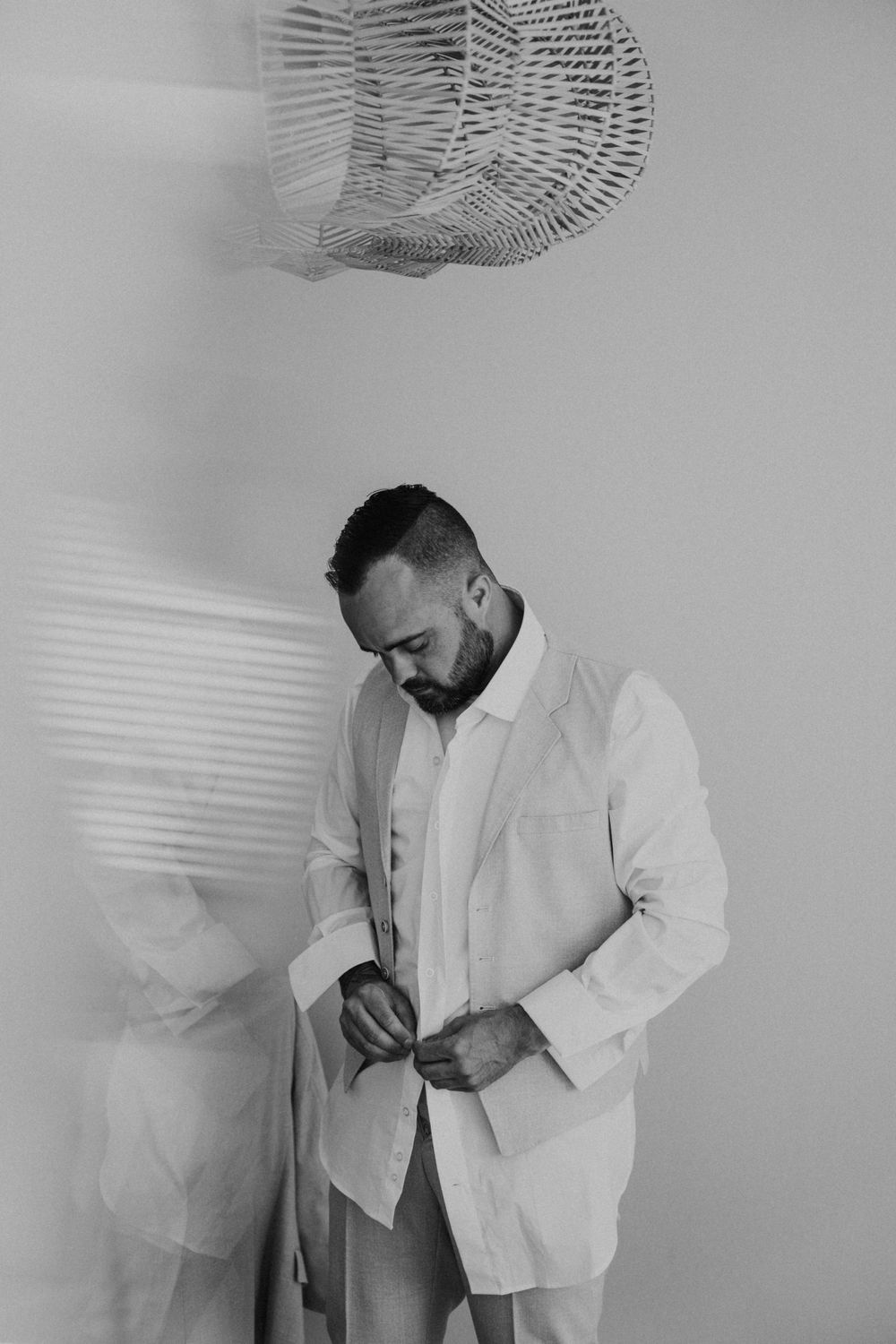 A person in a white suit jacket stands in a minimalist setting with a woven pendant lamp casting shadows above.