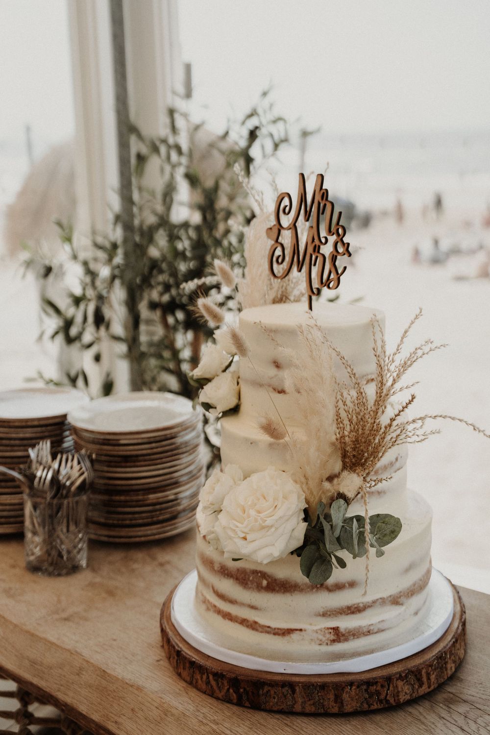 A rustic semi-naked wedding cake decorated with roses and dried pampas grass sits on a wooden slice next to stacked plates.