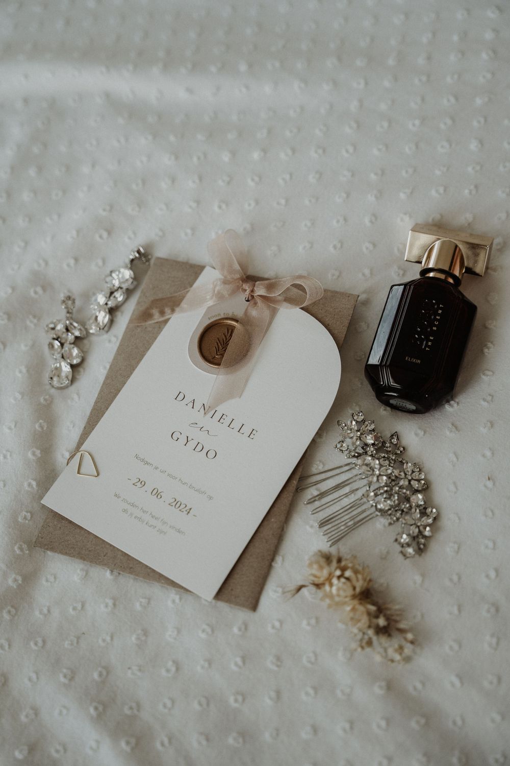 Wedding details flatlay with invitation, perfume bottle, crystal hair accessories and jewelry on textured white fabric.