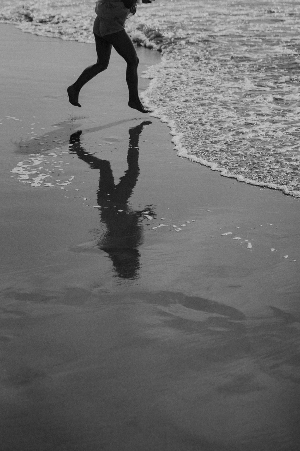 A silhouetted figure runs along the beach shoreline casting a reflection in the wet sand and incoming waves.