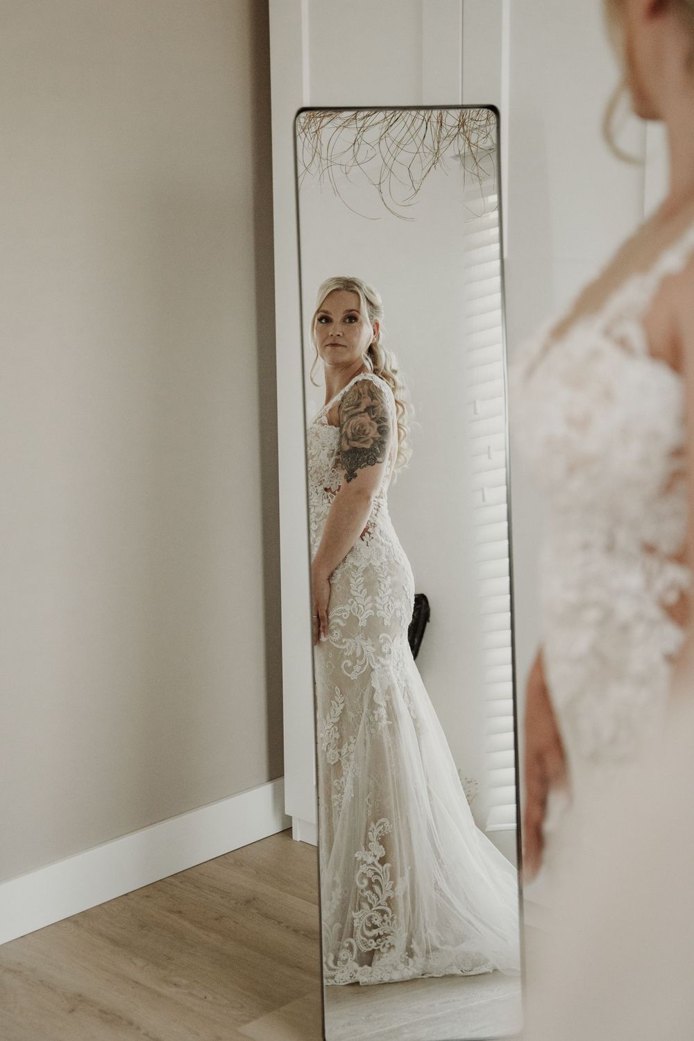 A bride wearing a detailed lace wedding gown with tattoos poses in front of a full-length mirror in a minimalist room.