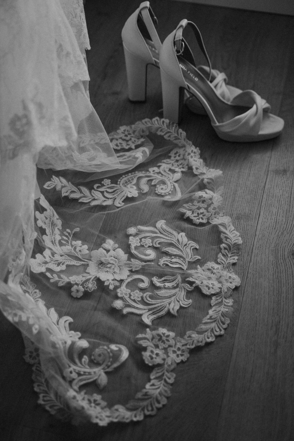 Black and white photo of delicate lace wedding veil detail and white high heel platform sandals on wooden floor.