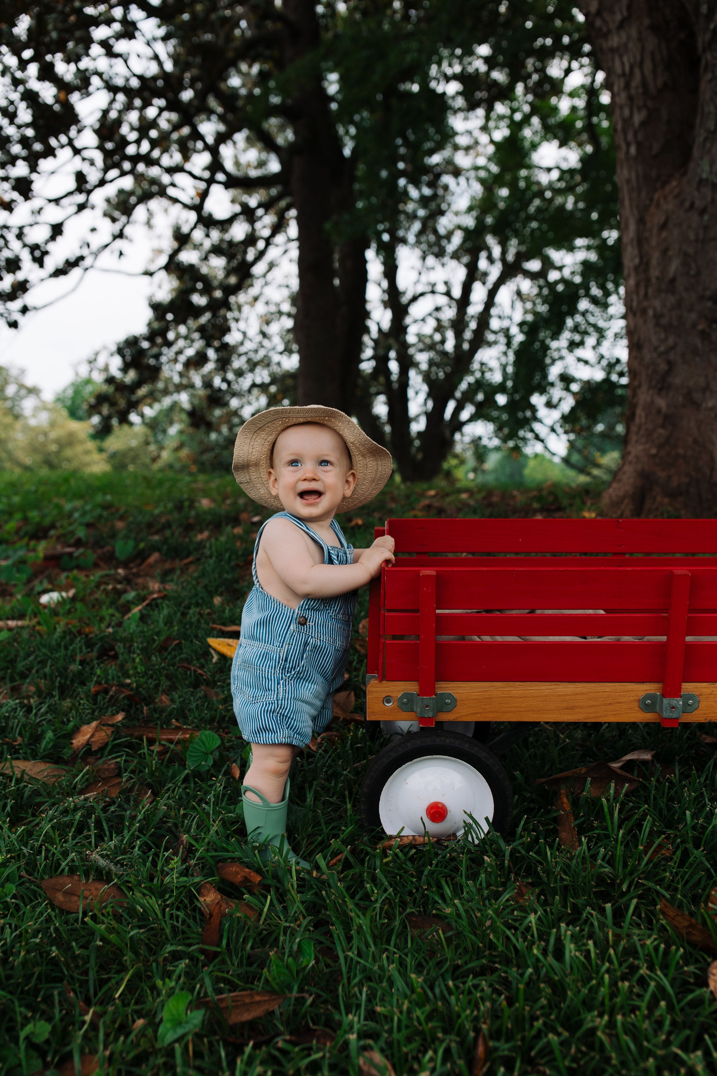 Child in overalls and boots standing next to red wagon outdoors.
