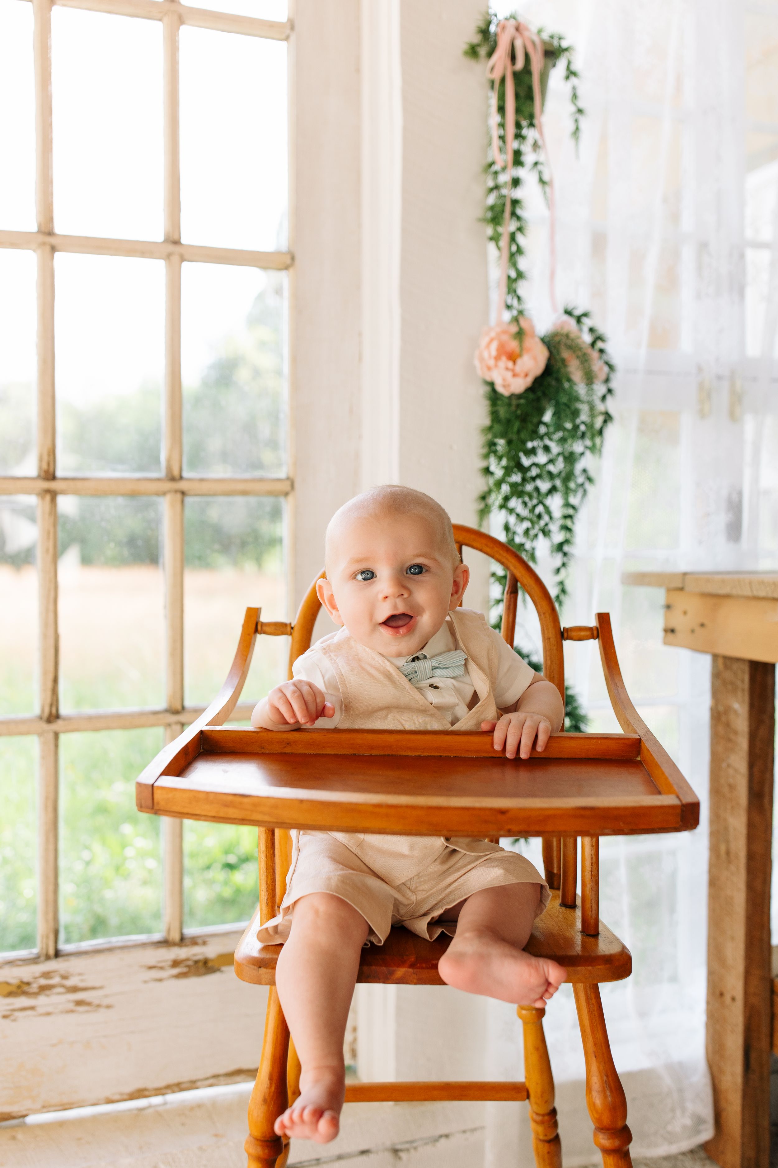 Baby in beige outfit sits in wooden high chair by sunny window with hanging plants.